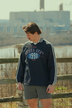 Person wearing a navy blue sweatshirt with 'Terra Ave 1990' print, standing by a wooden fence with a scenic background. travel and nature apparel