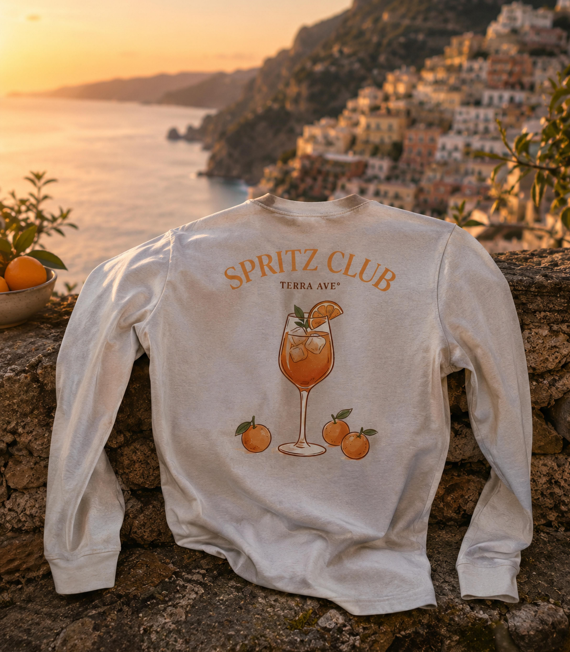 White sweatshirt with 'Spritz Club' design on a stone wall with a scenic amalfi coast background