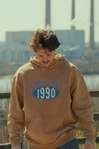 Person wearing a brown hoodie with 'Terra Ave 1990' text, standing outdoors with a cityscape background. travel style out in nature