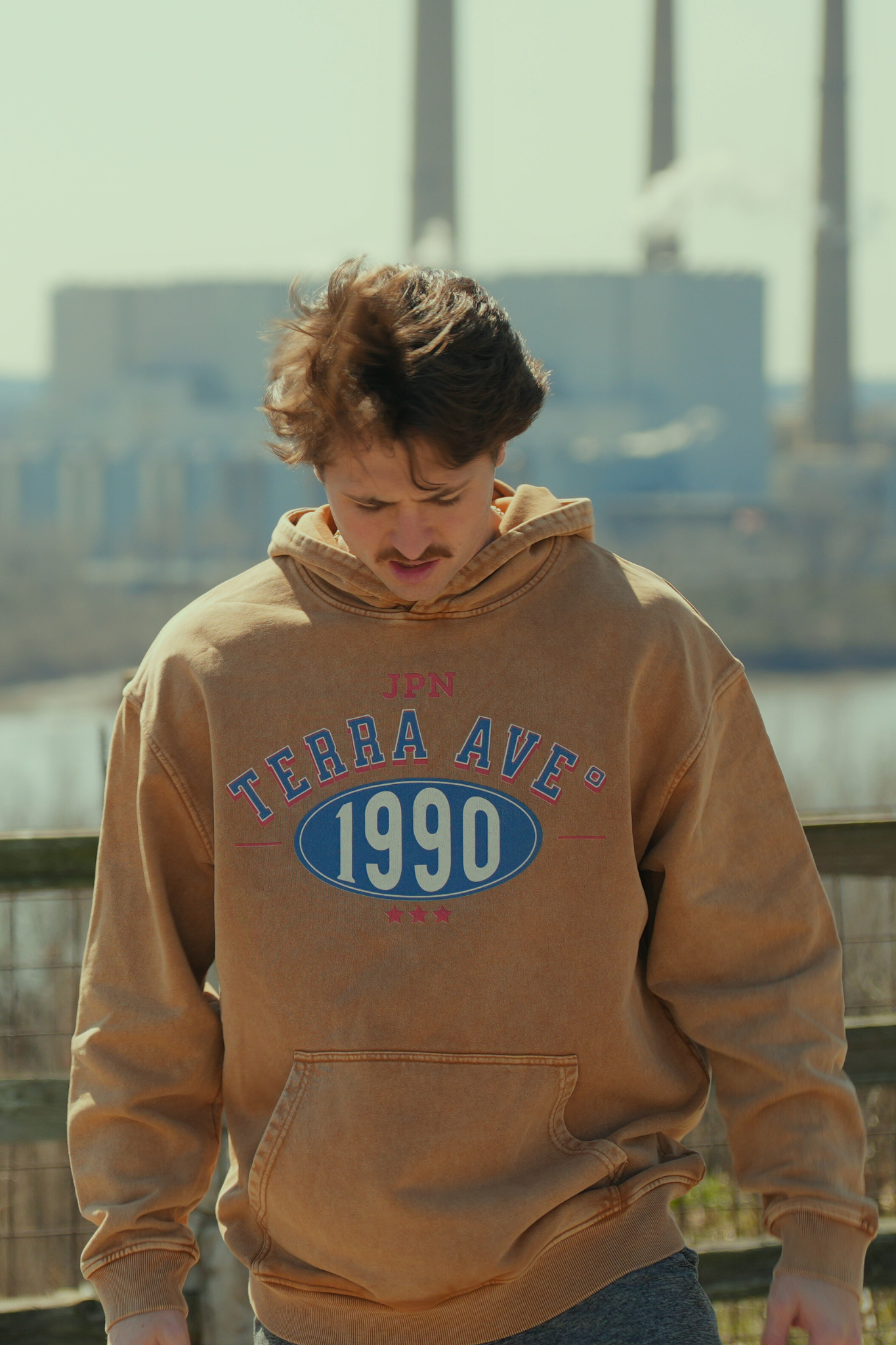 Person wearing a brown hoodie with 'Terra Ave 1990' text, standing outdoors with a cityscape background. travel style out in nature