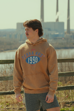Person wearing a brown hoodie with 'Terra Ave 1990' text, standing outdoors near a fence.