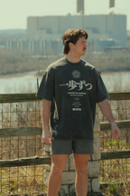 Person wearing a dark t-shirt with text and graphics, standing outdoors near a fence and industrial buildings. travel and nature clothing