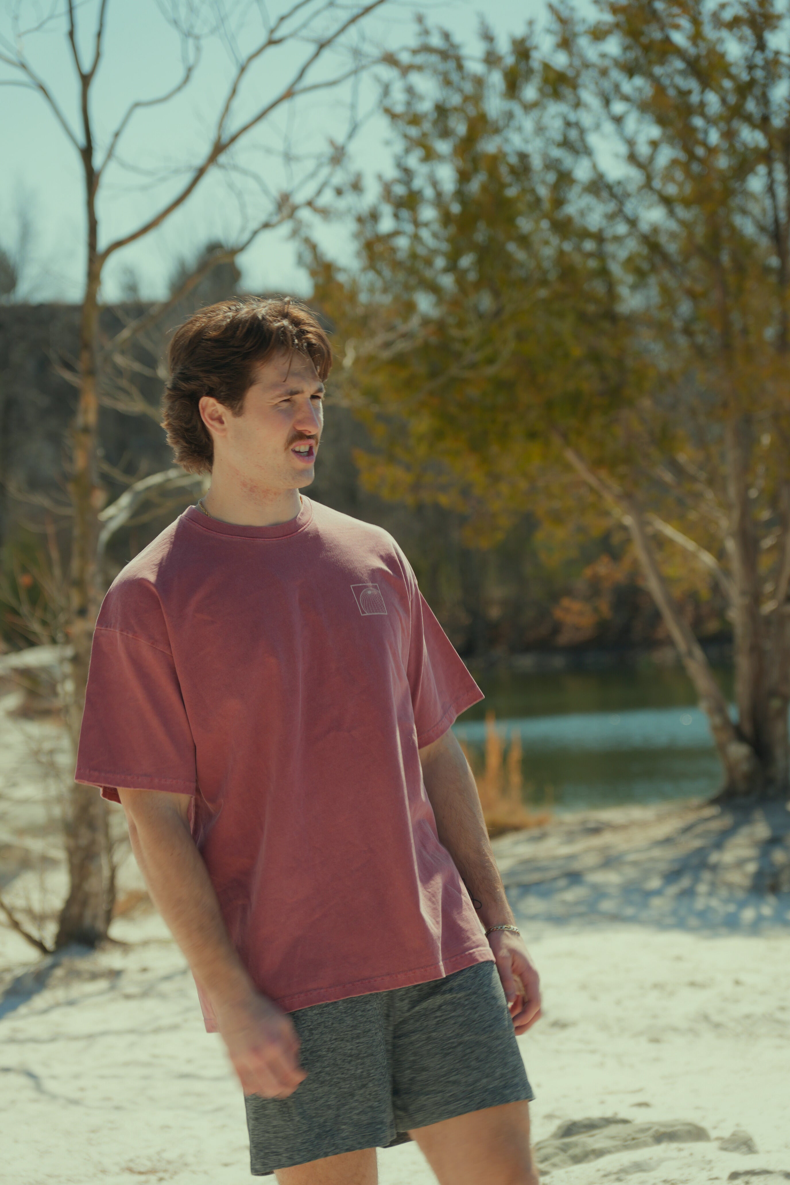 Man standing on a sandy beach with trees and water in the background wearing graphic travel tee