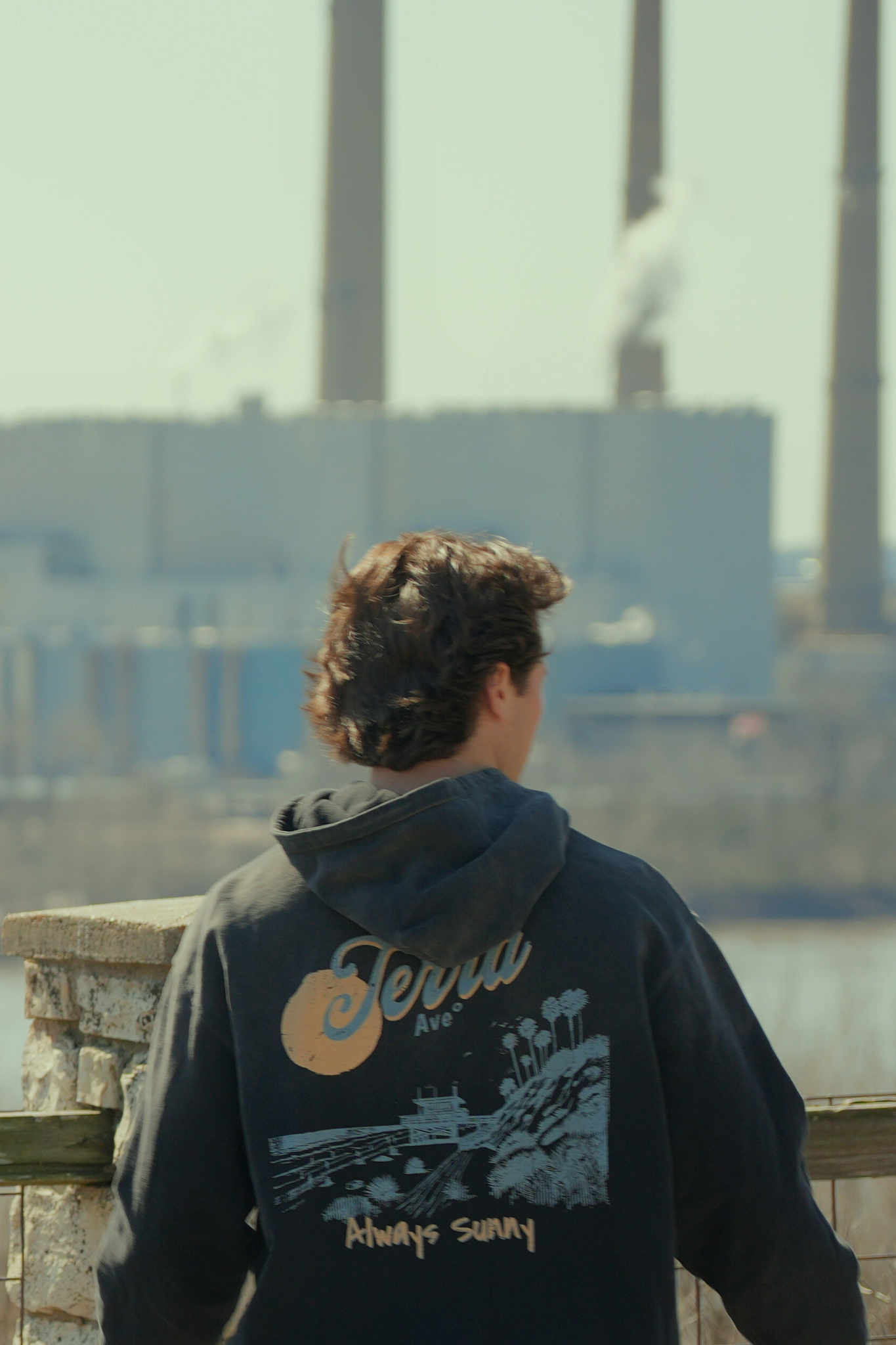 Person wearing a hoodie with 'Terra' and 'Always Sunny' text, standing by a waterfront with industrial buildings in the background. heavyweight faded hoodie
