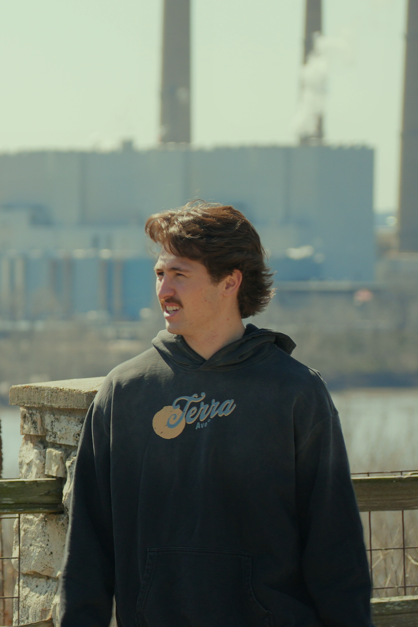 Man wearing a hoodie with a logo, standing by a waterfront with a blurred background heavyweight hoodie