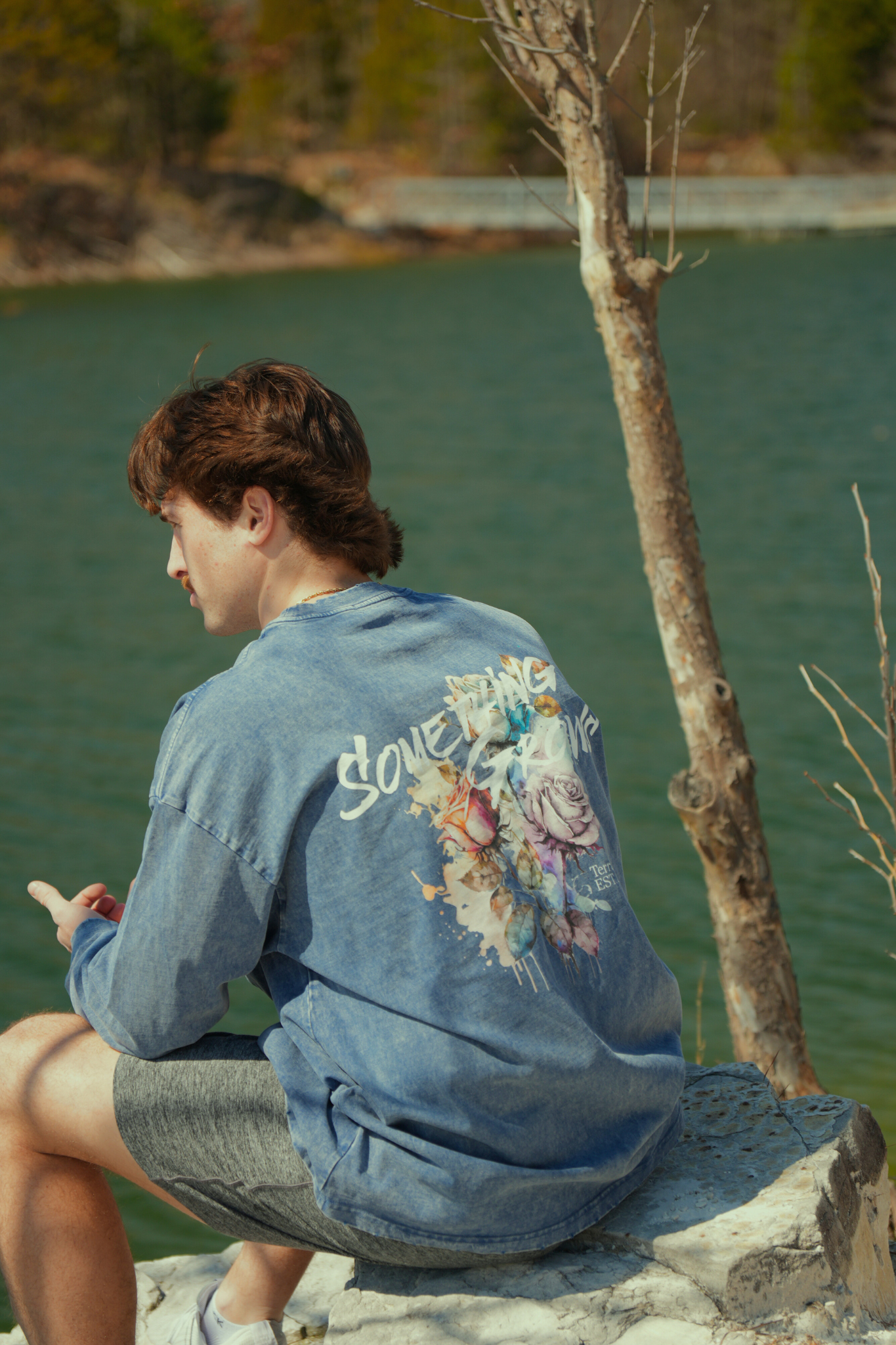 Person sitting by a lake wearing a denim long sleeve with floral designs. travel and nature 